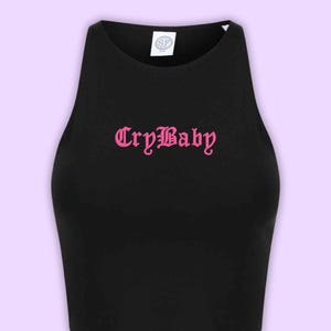 May include: A black sleeveless crop top featuring a high neckline and the text CryBaby printed in a pink gothic-style font across the chest. A white circular brand label with the letters SF is visible on the inside of the collar area.