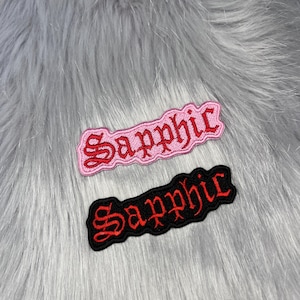 May include: Two embroidered patches featuring the word Sapphic in a gothic-style font. One patch has a light pink background with red lettering, and the other patch has a black background with red lettering. Both patches have a scalloped edge.