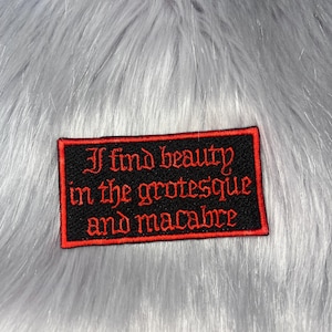 Peut inclure: Un écusson rectangulaire brodé avec un fond noir et une bordure rouge. Le texte "I find beauty in the grotesque and macabre" est cousu en fil rouge dans une police de style gothique au centre de la surface de l'écusson.