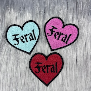 Puede incluir: Tres parches bordados para planchar en forma de corazón. Cada corazón es de un color diferente: azul claro, rosa y rojo. La palabra "Feral" está bordada en negro en cada corazón.