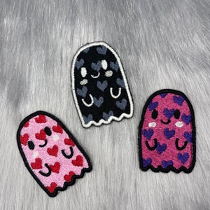 Könnte beinhalten: Drei gestickte Bügelbilder in Form von Geistern mit unterschiedlichen Hintergrundfarben. Der erste Geist ist schwarz mit grauen Herzen, der zweite ist rosa mit roten Herzen und der dritte ist lila mit blauen Herzen. Alle drei Geister haben weiße lächelnde Gesichter und weiße Herzen um die Augen.