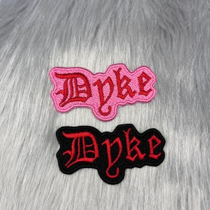 Könnte beinhalten: Zwei gestickte Aufnäher mit dem Wort Dyke in einer gotischen Schriftart. Ein Aufnäher hat eine rosa Schrift auf rosa Hintergrund, der andere eine rote Schrift auf schwarzem Hintergrund. Beide Aufnäher haben eine dem Text folgende Kontur.