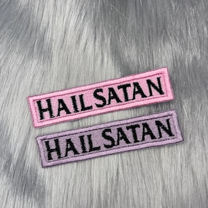 May include: Two embroidered patches with the text "Hail Satan" in black lettering. The top patch is pink and the bottom patch is purple.