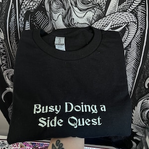 May include: Black t-shirt with the text "Busy Doing a Side Quest" embroidered in white on the front.