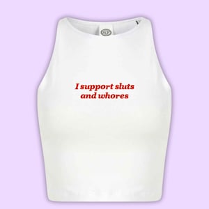 Op de afbeelding: Witte crop top met de rode tekst "I support sluts and whores". De top heeft een ronde hals en een klein logo bij de halslijn. De achtergrond is lichtpaars.