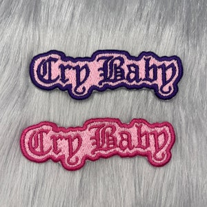 May include: Two embroidered patches with the text "Cry Baby" in a gothic font. The top patch is pink with purple stitching, and the bottom patch is purple with pink stitching.