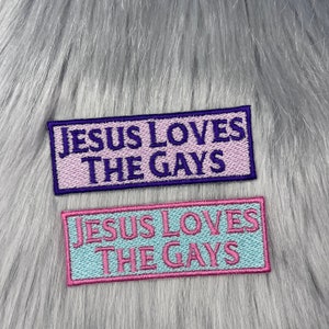 Peut inclure: Deux patchs brodés avec le texte "Jesus Loves The Gays" en lettres violettes et roses sur un fond bleu clair et rose.