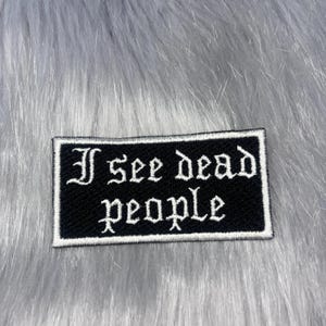 May include: A rectangular embroidered patch featuring a black background with a white border and the white text "I see dead people" written in a gothic-style font. The patch has a textured surface and is designed for application to fabric items.