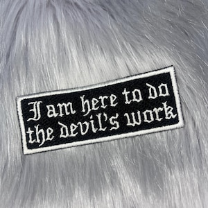 May include: A rectangular embroidered patch with a black background and a white border. The patch features the white text "I am here to do the devil's work" written in a gothic-style font, centered across two lines on the fabric surface.
