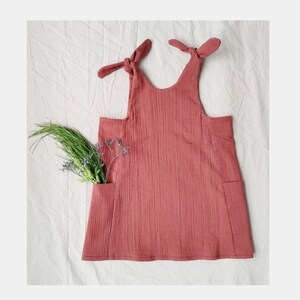 May include: A dusty rose dress with tie straps and two front pockets. The dress is made of a textured fabric and has a relaxed fit. A sprig of green and purple flowers is tucked into one of the pockets.