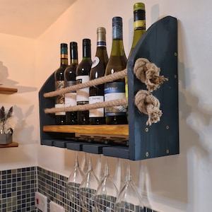 May include: A dark blue wooden wine rack with six bottles of wine and six wine glasses. The rack is mounted on a wall and has a rope design.