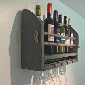 May include: A dark gray wooden wall-mounted wine rack with rope accents. The rack holds six bottles of wine and has four glass holders underneath.