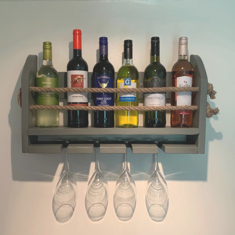 May include: A gray wooden wine rack with rope accents holds six bottles of wine and has four glass stemware hanging below. The bottles are labeled "Vibrant Gravity", "Troubadour", "Toro Loco", "G", "Pinotage", and "Gallo".