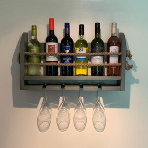 May include: A gray wooden wall-mounted wine rack with six bottles of wine and six wine glasses hanging below. The rack is decorated with rope and has a rustic look.