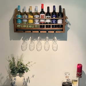 Może przedstawiać: Drewniany stojak na wino z siedmioma butelkami wina i sześcioma kieliszkami do wina wiszącymi na stojaku. Stojak jest zamontowany na białej ścianie. Butelki to mieszanka wina czerwonego i białego. Kieliszki są przezroczyste i mają n&oacute;żkę.