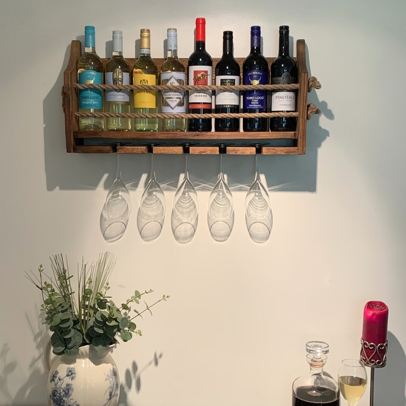 Może przedstawiać: Drewniany stojak na wino z siedmioma butelkami wina i sześcioma kieliszkami do wina wiszącymi na stojaku. Stojak jest zamontowany na białej ścianie. Butelki to mieszanka wina czerwonego i białego. Kieliszki są przezroczyste i mają n&oacute;żkę.