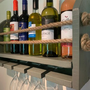 May include: A rustic wooden wine rack with a gray paint finish and rope accents. The rack holds six bottles of wine and has six glass holders underneath. The bottles are a mix of red and white wine.