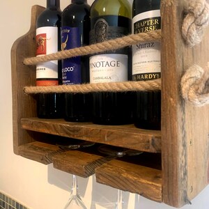 May include: A rustic wooden wine rack with rope accents holds five bottles of wine and has three glass holders underneath. The bottles are labeled "Oloco", "Pinotage", "Chapter & Verse", and "Hardys".
