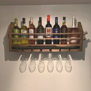 May include: A rustic wooden wine rack with rope accents holds eight bottles of wine and six wine glasses. The rack is mounted on a white wall.
