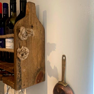 May include: A rustic wooden wine rack with rope accents and a copper pan hanging on the wall behind it. The rack has a shelf for bottles and a lower shelf for glasses.