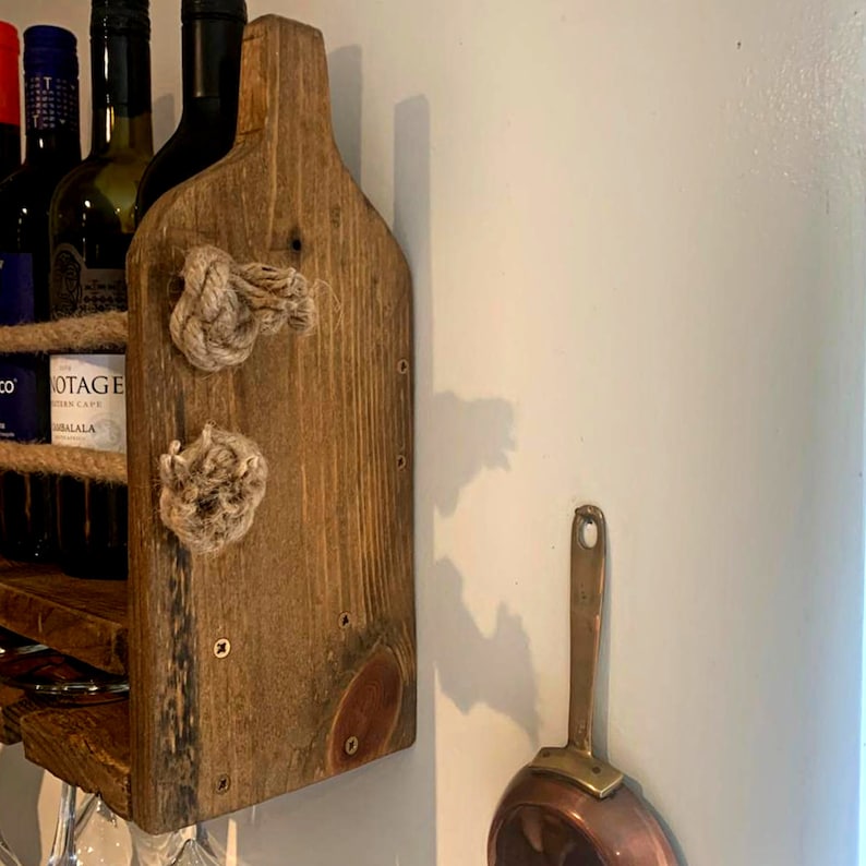 May include: A rustic wooden wine rack with rope accents and a copper pan hanging on the wall behind it. The rack has a shelf for bottles and a lower shelf for glasses.