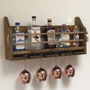 May include: A rustic wooden wall-mounted shelf with rope accents, holding four glass bottles of liquor and four copper-coloured wine glasses. The bottles are labelled "Little Bird", "The Fife", "Gunpowder", and "Mr. Soul Gin".