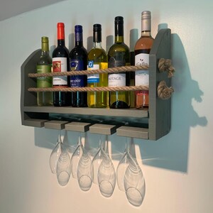May include: A rustic wooden wall-mounted wine rack with a green finish. The rack has two shelves and a rope design. It holds six bottles of wine and has four glass holders underneath.