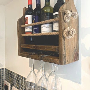 May include: Rustic wooden wine rack with rope accents, featuring four bottles of wine and three wine glasses hanging below. The rack is mounted on a white wall.