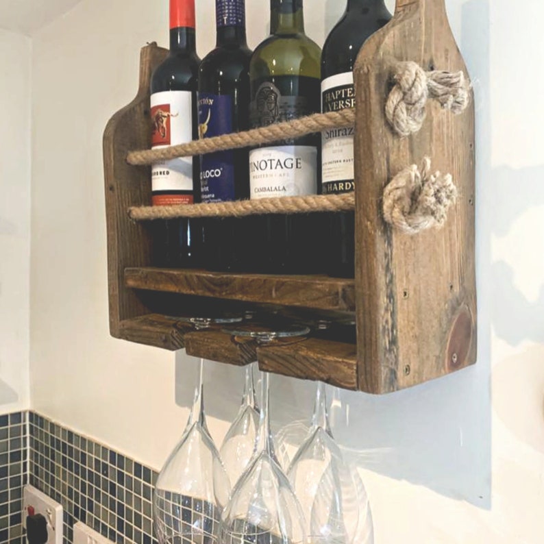 May include: Rustic wooden wine rack with rope accents, featuring four bottles of wine and three wine glasses hanging below. The rack is mounted on a white wall.