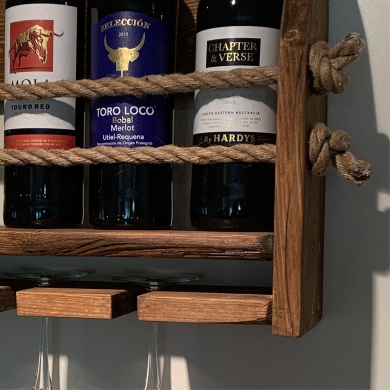 Może przedstawiać: Drewniany stojak na wino z akcentami z liny, na kt&oacute;rym znajdują się trzy butelki wina. Butelki są oznaczone etykietami "Touro Red", "Selecci&oacute;n 2018" i "Chapter & Verse by Hardys". Stojak ma r&oacute;wnież dwa uchwyty na kieliszki.