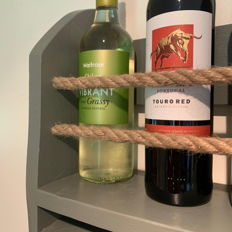 May include: A gray wooden wine rack with two bottles of wine. The wine rack has two shelves and is decorated with rope. The bottle on the left is a white wine labeled "Waitrose Chilean Vibrant and Grassy Valle Central". The bottle on the right is a red wine labeled "Portugal Touro Red Edi&ccedil;&atilde;o Limitada".