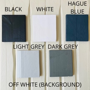 May include: A collection of paint color samples, including black, white, light grey, dark grey, off white, and blue. The samples are arranged in a row on a white wooden surface.
