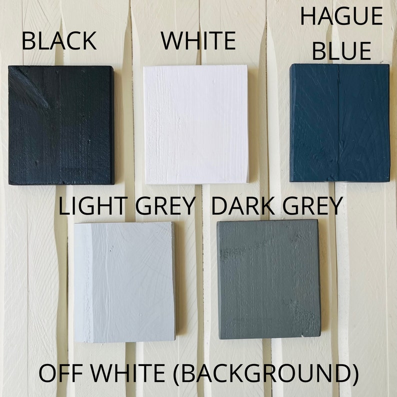 May include: A collection of paint color samples, including black, white, light grey, dark grey, off white, and blue. The samples are arranged in a row on a white wooden surface.