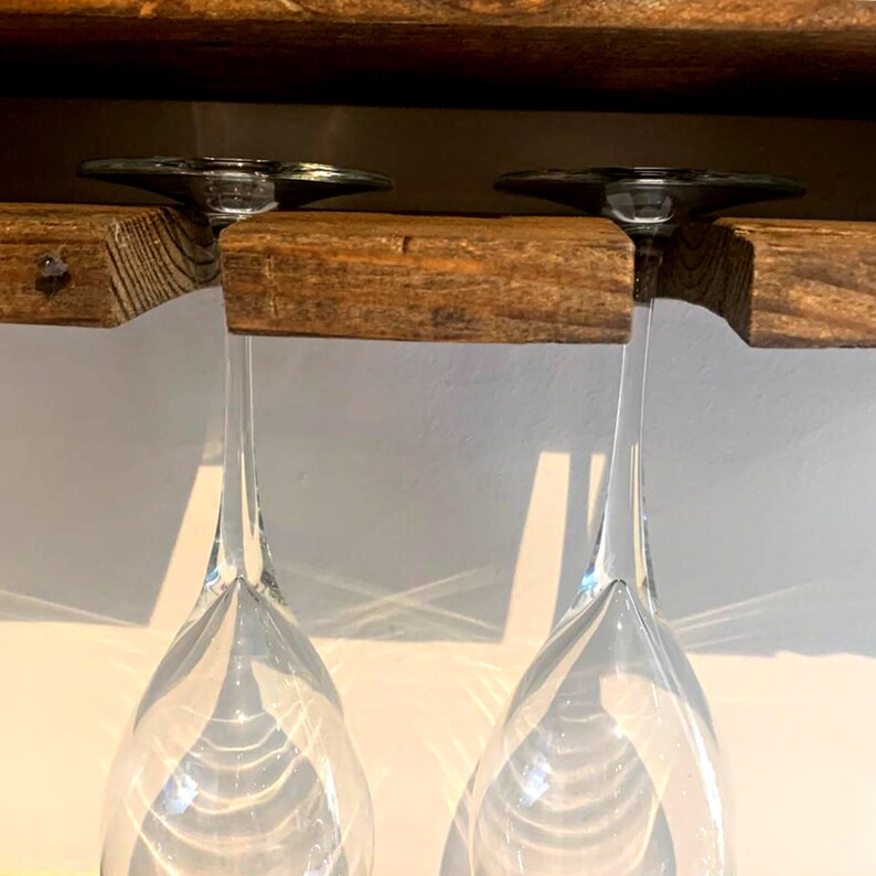 May include: Two clear glass wine glasses hang upside down from a wooden shelf. The shelf is made of reclaimed wood and has a rustic look.