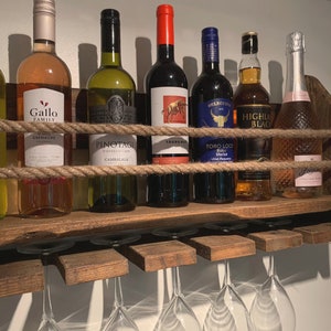 May include: A wooden wine rack with rope accents holds various bottles of wine and spirits, including Gallo Family Grenache, Pinotage, Tourored, Belleccio Toro Loco Bobal Merlot, Highland Black Blended Scotch, and a bottle of Italian Prosecco. The rack has six glass holders underneath.