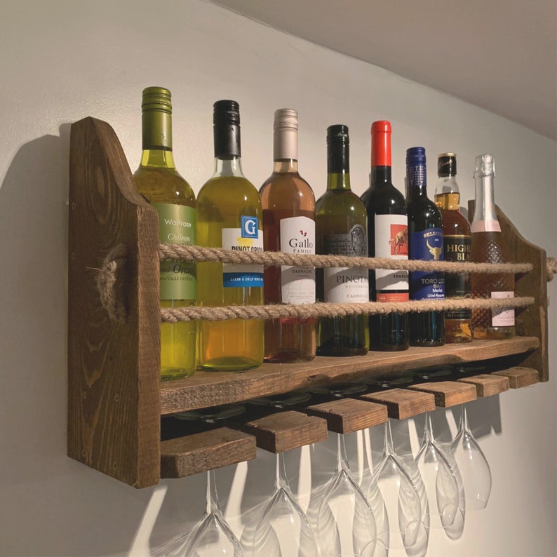 May include: A wooden wine rack with a rope design holds eight bottles of wine. The rack is mounted on a wall and has six wine glasses hanging from it. The bottles are a mix of red and white wine.