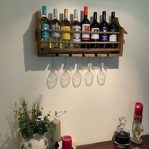 Może przedstawiać: Drewniany stojak na wino montowany na ścianie z detalem z sznurka, kt&oacute;ry mieści osiem butelek wina i sześć kieliszk&oacute;w do wina. Stojak jest w kolorze ciemnobrązowym i jest zamontowany na białej ścianie.