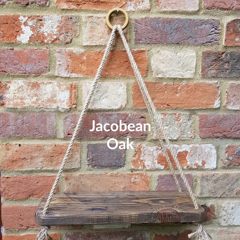 May include: A wooden shelf with a Jacobean Oak finish, hanging from a brick wall with natural rope. The shelf is suspended from a gold metal ring.