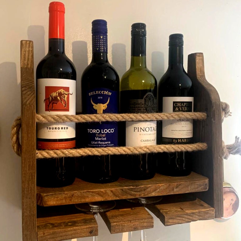 May include: A rustic wooden wine rack with rope accents holds four bottles of wine and has a shelf for glasses. The bottles are labeled "Touro Red", "Toro Loco Bobal Merlot", "Pinot Noir", and "Chapte & Versenne".