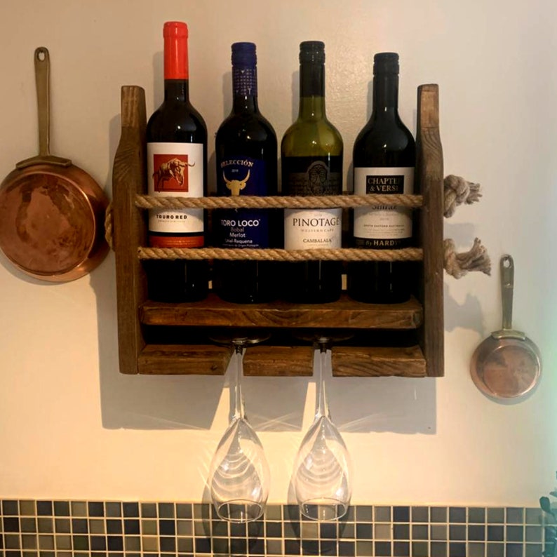 May include: A rustic wooden wine rack with rope accents, holding four bottles of wine and two wine glasses. The rack is mounted on a white wall with a tiled backsplash.
