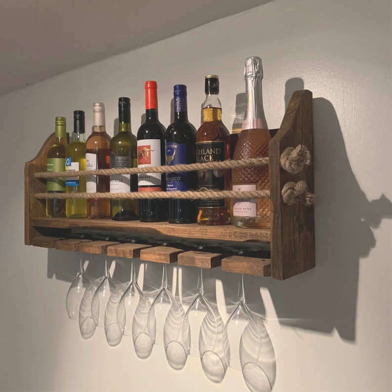 May include: A rustic wooden wine rack with a rope design, holding various bottles of wine and spirits. The rack has a built-in glass holder underneath.