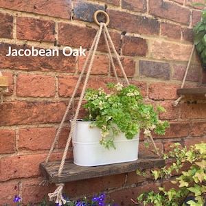 May include: A wooden shelf with a hanging planter made of rope. The shelf is stained in a Jacobean Oak finish. A white metal planter with green plants hangs from the rope. The shelf is mounted on a brick wall.