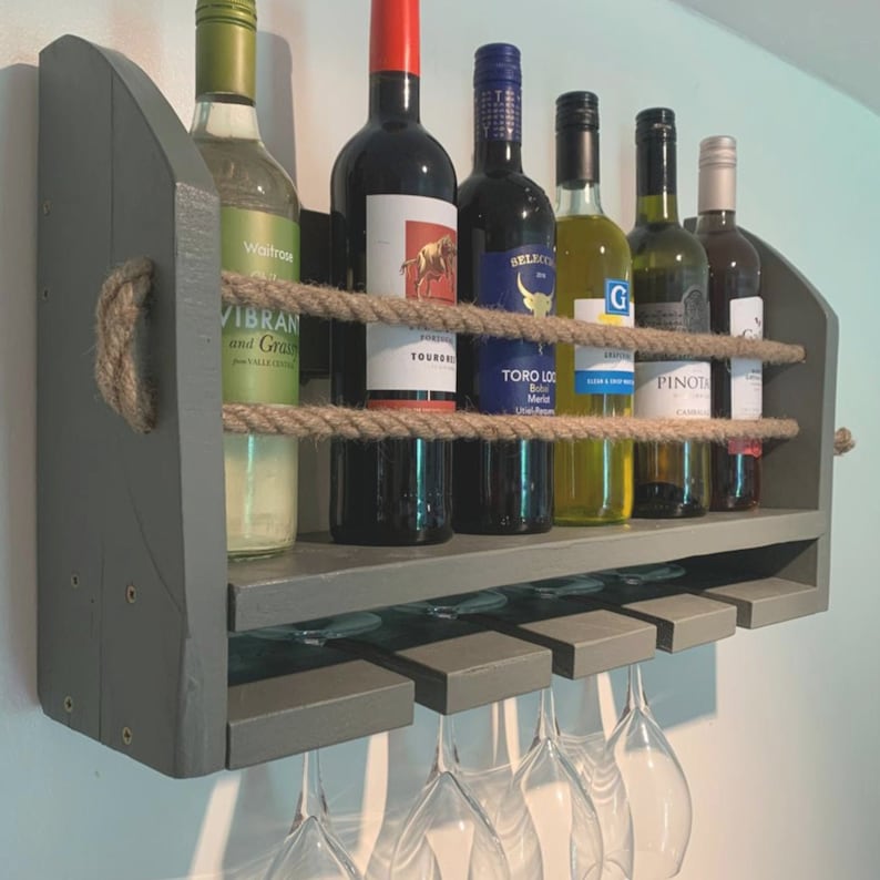 May include: A gray wooden wall-mounted wine rack with rope accents. The rack holds six bottles of wine and has six glass holders underneath. The bottles are labeled with various names, including "Toro Loco" and "Pinot Noir".