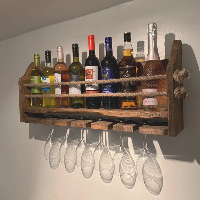 May include: A rustic wooden wine rack with a rope design, holding eight bottles of wine and a set of eight wine glasses. The rack is mounted on a white wall.