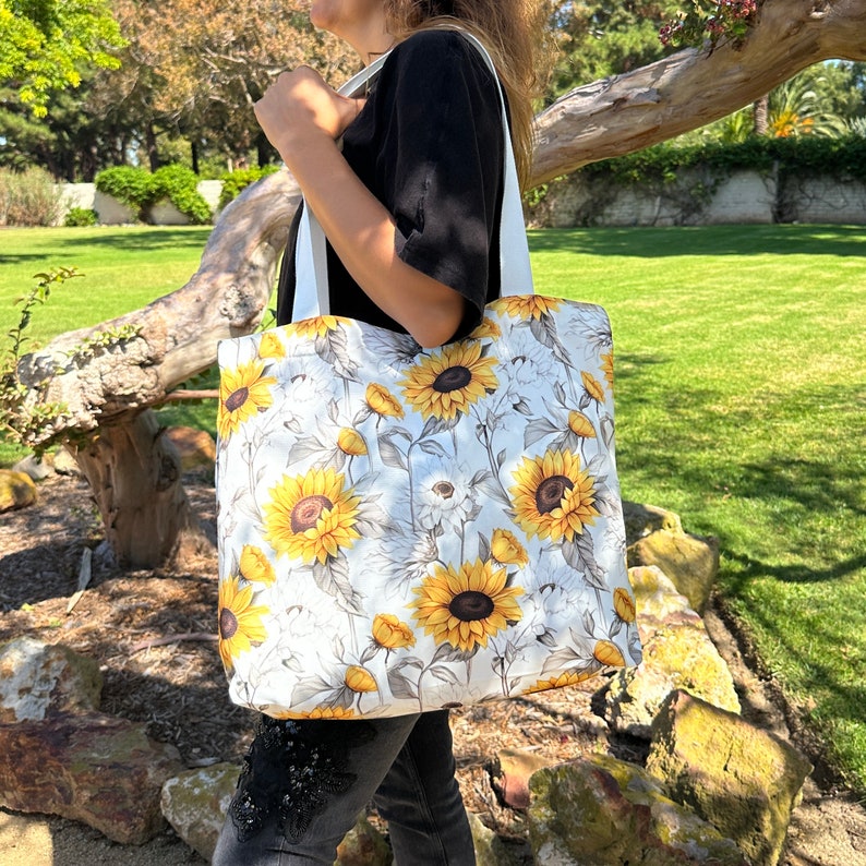 K&ouml;nnte beinhalten: Eine wei&szlig;e Canvas-Tragetasche mit einem Sonnenblumenmuster. Die Tasche hat wei&szlig;e Henkel und wird von einer Person getragen.