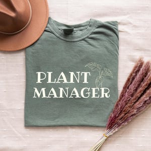 Può includere: T-shirt verde salvia con le parole "PLANT MANAGER" in lettere bianche e un'illustrazione di una pianta. Un cappello marrone e un mazzo di pampas rosa secche sono anche nell'immagine.