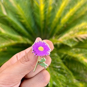 May include: A gold-tone enamel pin featuring a purple flower with a yellow center and green leaves.