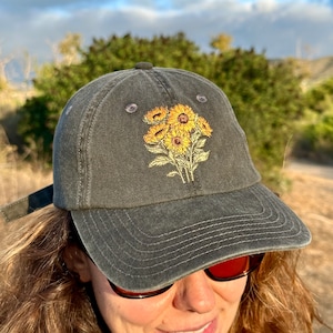May include: A dark gray baseball cap with an embroidered design of yellow sunflowers. The cap has a curved brim and a strap in the back.