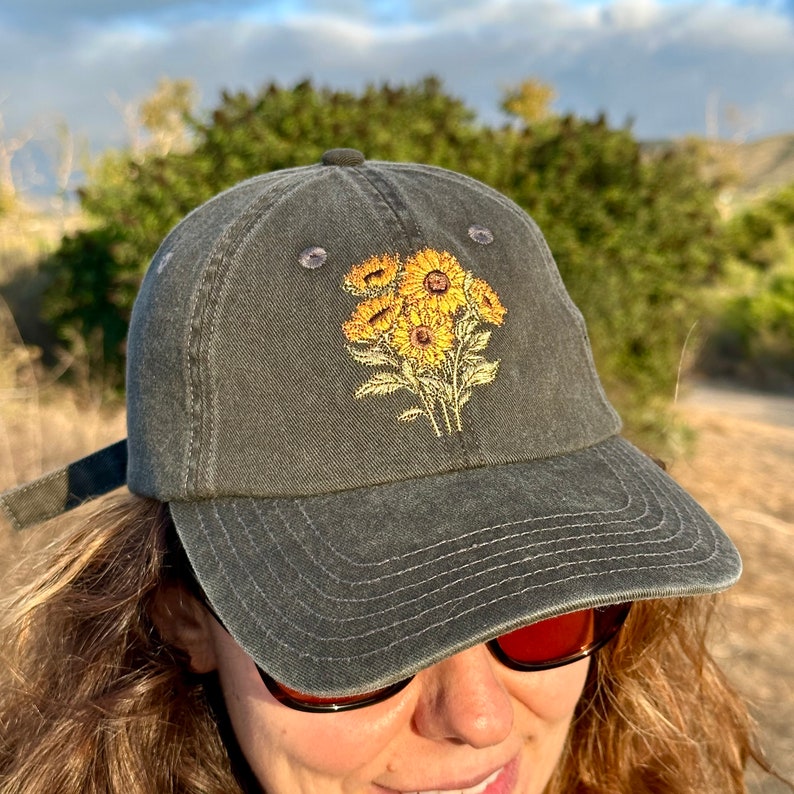 May include: A dark gray baseball cap with an embroidered design of yellow sunflowers. The cap has a curved brim and a strap in the back.