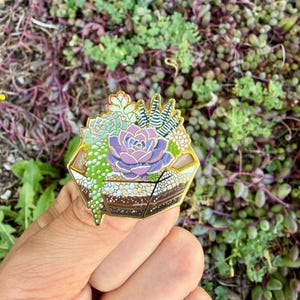 May include: A gold-coloured enamel pin depicting a terrarium with succulents. The pin features a central purple succulent surrounded by green and white plants. The terrarium has a layered brown and white base.